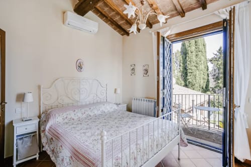 Bright bedroom at Casa Rossi Roma with white metal bed, exposed wooden beams, and French doors opening to terrace with