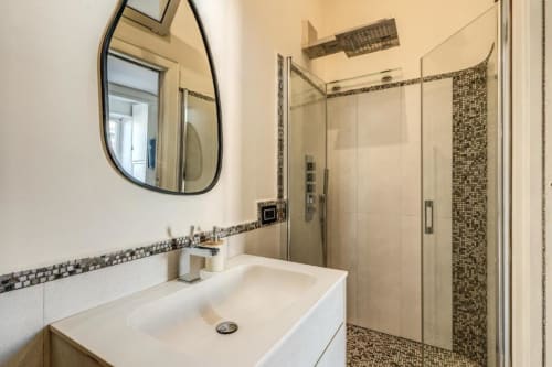 Bagno moderno in appartamento romano con lavabo bianco, specchio ovale, cabina doccia in vetro e dettagli in piastrelle mosaico