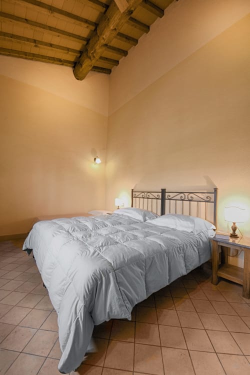 Camera da letto rustica a Villa Olena con biancheria bianca, letto in ferro battuto, travi in legno a vista e pavimento in cotto