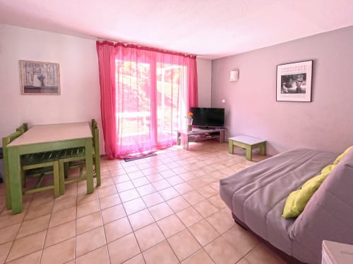 Studio rose à Digne-les-Bains avec canapé, table à manger, TV et porte-fenêtre avec rideaux rose