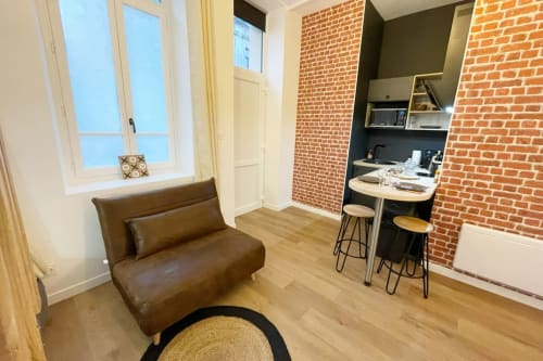 Studio moderne Les Logis d'Emma : canapé marron, kitchenette briques apparentes, parquet, vue sur la ville.
