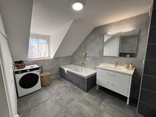 Salle de bain moderne du grenier aux Logis d'Emma avec baignoire, vasque blanche, miroir et lave-linge en France
