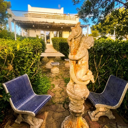Cortile di lusso a My Home Apulia con scultura ornamentale di sirena in pietra, sedie blu e siepi curate
