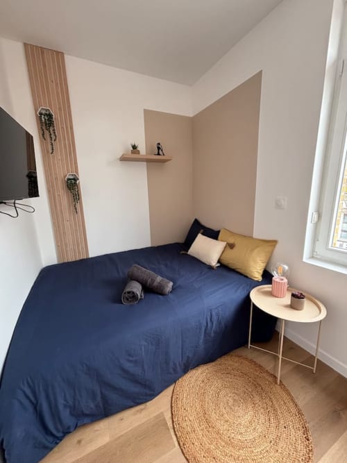 Chambre cosy aux Logis d'Emma avec lit bleu marine, coussins moutarde, mur en bois et tapis jute rond.