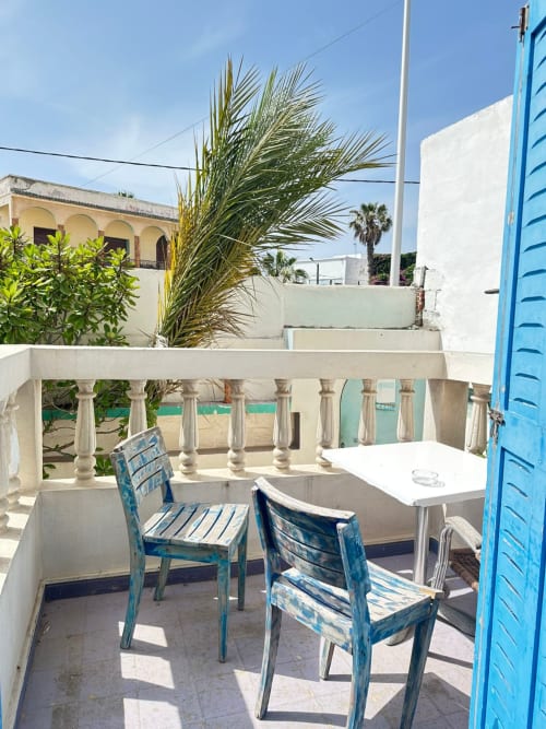 Sunny terrace at Yousurf Surfhouse hostel in Essaouira with blue chairs, white table, and palm tree overlooking beach