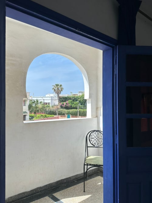 Circular window in Yousurf Surfhouse Berber room, Essaouira, viewing lush garden with palm tree and blue sky