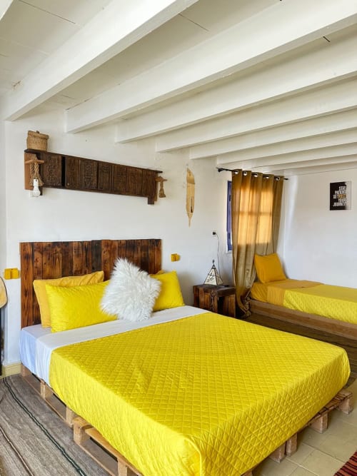 Bright yellow bedding in Sahara room at Yousurf Surfhouse hostel, Essaouira, with wooden headboard and white beam ceiling