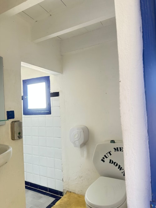 Bathroom in Yousurf Surfhouse hostel, Essaouira with white tiles, toilet with humor sign, and blue window