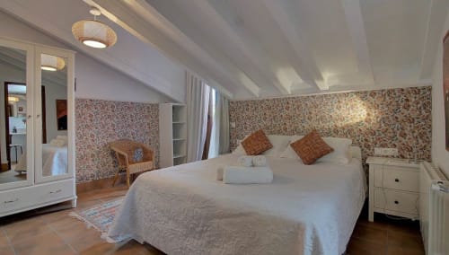 Dormitorio luminoso en Casa Gallinera en Benissivà con papel pintado floral, ropa de cama blanca, vigas de madera y vistas a las montañas