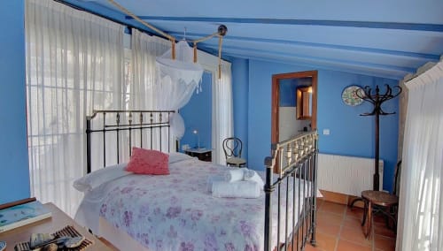 Dormitorio azul brillante con cama de hierro forjado, cortinas blancas translúcidas y vistas a la montaña en Casa Gallinera