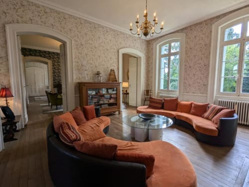 Élégant salon du Domaine du Faure avec sièges orange, lustre, cheminée et vue sur les jardins par de hautes fenêtres.