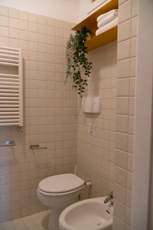 Bagno moderno dell'A-waay Hotel Torino con rivestimenti in piastrelle color crema, toilet, bidet, portasciugamani e pianta verde cascante
