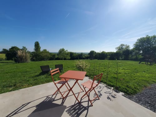 Salon de jardin en terrasse du gîte Le Clos Des Sources à Isigny-sur-Mer, table et chaises rouges avec vue sur le jardin verdoyant