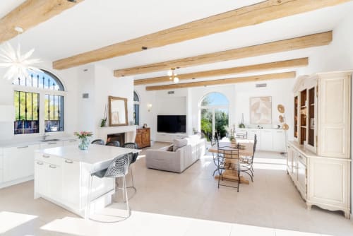 Bright open-plan villa interior with exposed wooden beams, white kitchen island, seating area, fireplace, and arched windows