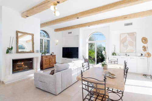 Bright modern villa living room with fireplace, white seating, wooden beams, and arched doors opening to pool view, Cannes