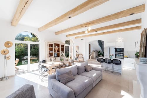 Modern villa living area with grey sofa, exposed beams, kitchen, dining table, and garden view through arched doors in Cannes