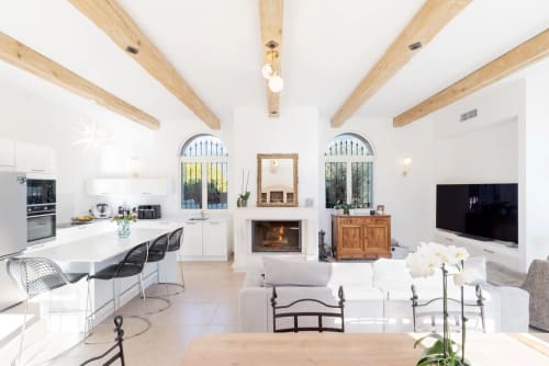 Modern open-plan villa living area with exposed wooden beams, fireplace, white furnishings, and integrated kitchen in