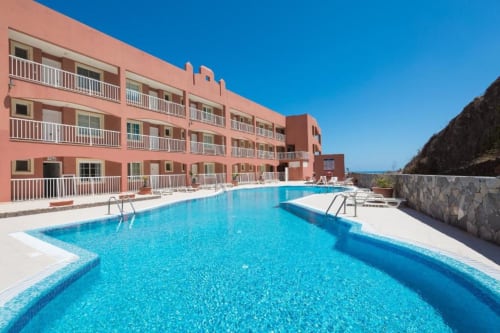 Vista luminosa de la piscina de Smart Rent Fuerteventura con edificio coral rosado, piscina azul cristalina y cielo despejado en La