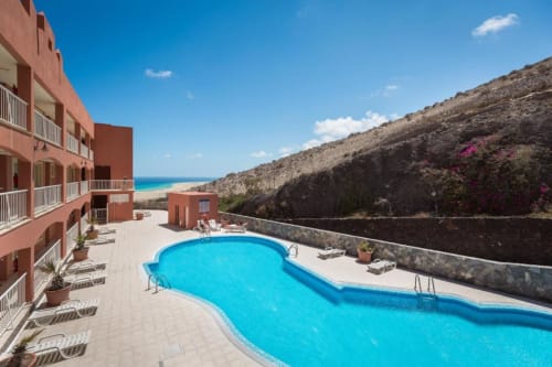 Piscina de Smart Rent Fuerteventura con terraza, edificio rojo, playa y acantilados al fondo