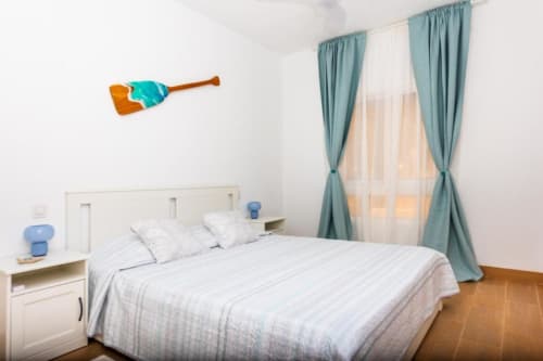 Dormitorio luminoso en apartamento Brisa del Atlántico con cama queen, ropa de cama blanca, cortinas turquesas y piso de madera