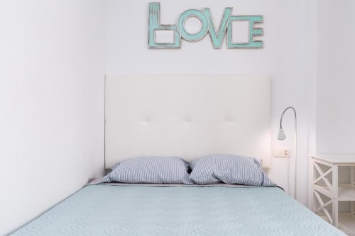 Habitación luminosa en Smart Rent Fuerteventura con letrero HOME turquesa, cabecero blanco y almohadas a rayas