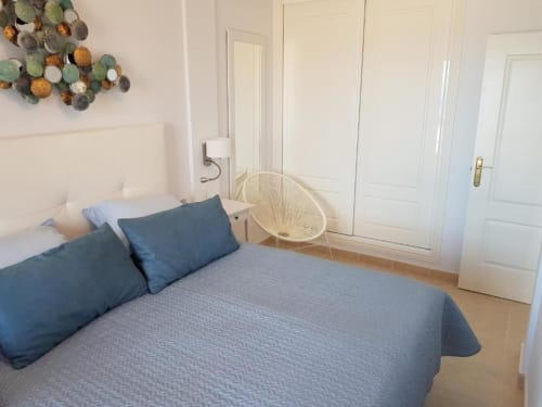 Luminoso dormitorio en Smart Rent Fuerteventura con ropa de cama azul, cabecero blanco, sillón crema y armario blanco