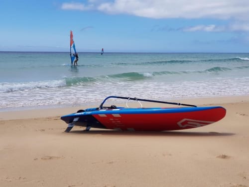 Tabla de windsurf roja y azul en playa arenosa con windsurfistas en olas del océano en alquiler vacacional de Fuerteventura