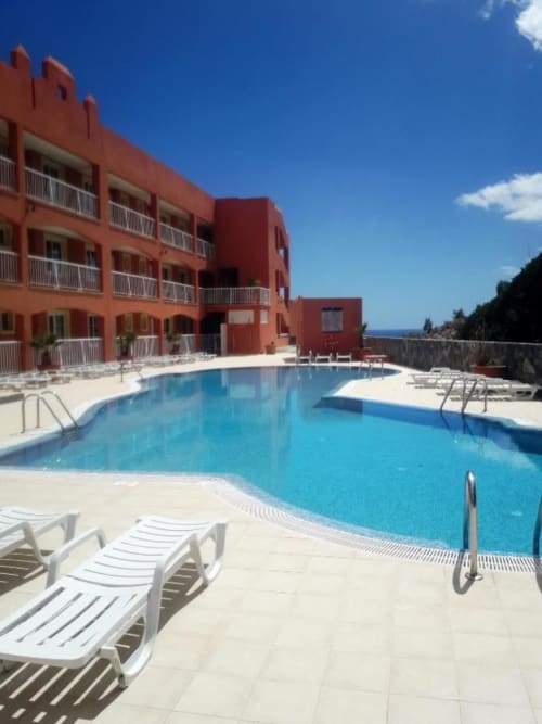 Edificio rojo de resort con piscina azul brillante, tumbonas y vista al océano en Smart Rent Fuerteventura