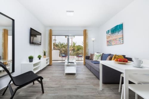 Moderno salón de apartamento en Smart Rent Fuerteventura con sofá, zona de comedor, cocina y vistas al jardín desde las puertas del patio