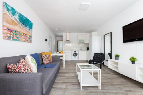 Apartamento moderno de planta abierta en Smart Rent Fuerteventura con sofá gris, cocina blanca, comedor y arte de playa