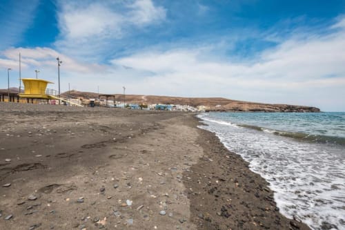 Playa de Tarajalejo con torre de socorrista amarilla, arena negra, olas y edificios costeros bajo cielo azul en Fuerteventura