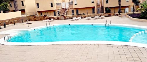 Piscina curva de agua turquesa rodeada de tumbonas blancas en el complejo de apartamentos Smart Rent Fuerteventura con terraza y