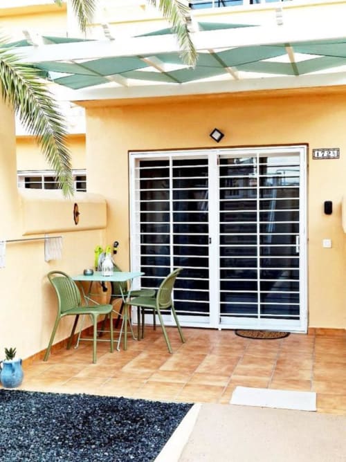 Terraza de la Casa de Alex en Smart Rent Fuerteventura con paredes amarillas, sillas verdes, mesa de comedor y vistas al jardín bajo pérgola