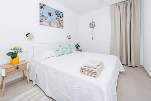 Dormitorio luminoso en Smart Rent Fuerteventura con cama blanca, almohadas azules, mesita de noche de madera con flores amarillas y tonos beige