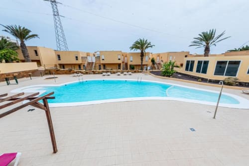 Piscina exterior luminosa con tumbonas en el complejo de apartamentos Smart Rent Fuerteventura, con edificios amarillos y palmeras