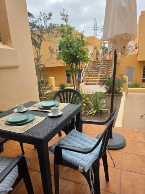 Terraza de comedor al aire libre en Smart Rent Fuerteventura con mesa preparada, sillas negras y edificios amarillos con vistas al jardín