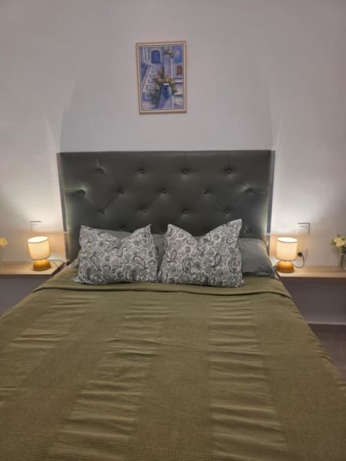 Moderno dormitorio en Smart Rent Fuerteventura con cabecero capitonado gris, ropa de cama verde oliva, almohadas estampadas y lámparas de noche