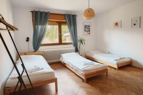 Bright bedroom with three wooden single beds, white linens, parquet floors, window with teal curtains overlooking mountains