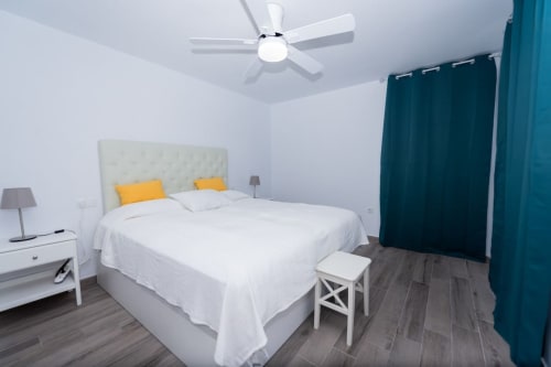 Dormitorio moderno en Casa Andre con cama blanca, cortinas turquesas, ventilador de techo y piso de madera oscura en Fuerteventura