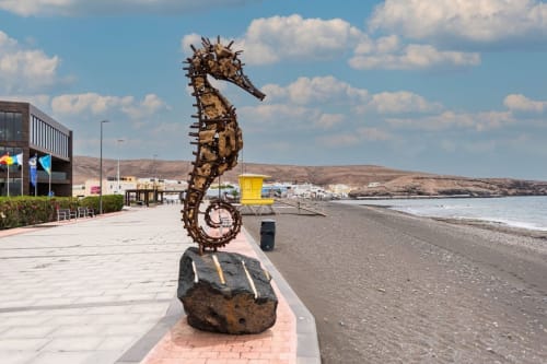Escultura de caballito de mar en el paseo marítimo de Fuerteventura con pueblo costero y montañas al fondo