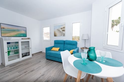 Moderno salón con sofá turquesa, mesa de comedor blanca, decoración costera y ventanales con vistas al océano en Smart Rent