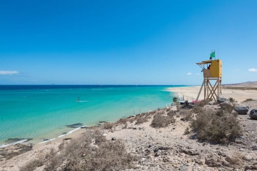 Playa turquesa con torre de vigilancia amarilla en apartamento Smart Rent Fuerteventura, cielo azul despejado