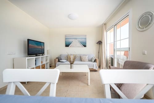 Luminoso salón con sofá, televisión, sillas blancas de comedor y acceso a balcón con vistas a la playa en Smart Rent