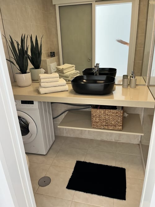 Cuarto de baño moderno con lavabos negros de cerámica, toallas crema, plantas en macetas y lavadora en Smart Rent Fuerteventura