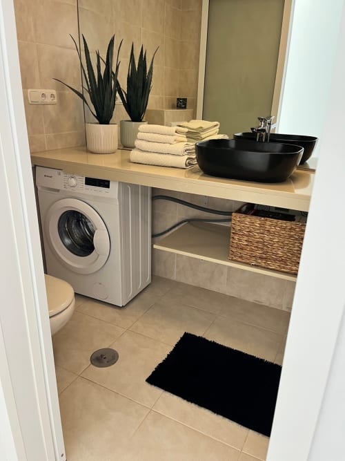 Moderna lavandería en apartamento Smart Rent Fuerteventura con lavadora, lavamanos negro, plantas decorativas y toallas dobladas