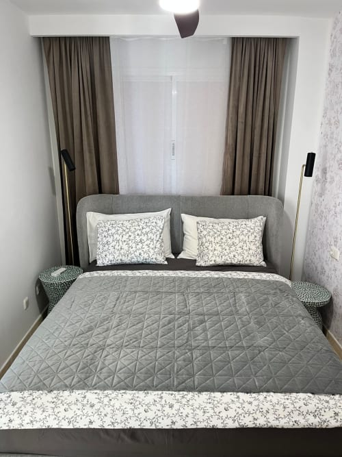 Moderno dormitorio de apartamento Smart Rent Fuerteventura con cama tapizada gris, almohadas florales y cortinas topo
