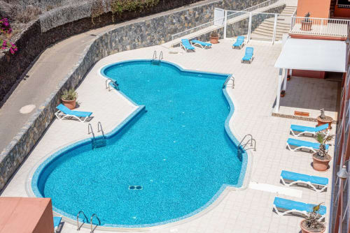 Vista aérea de la piscina en forma de riñón de Smart Rent Fuerteventura con tumbonas azules, terraza y muros de piedra en España.
