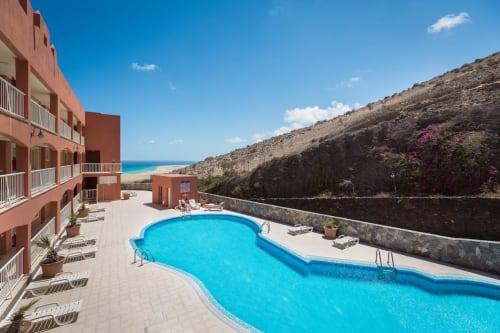 Resort Smart Rent Fuerteventura con piscina azul, edificios de terracota roja, acceso a playa y paisaje volcánico en Canarias