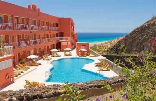 Hotel rojo frente al mar con piscina curva azul, sombrillas blancas y vista a la playa en Fuerteventura