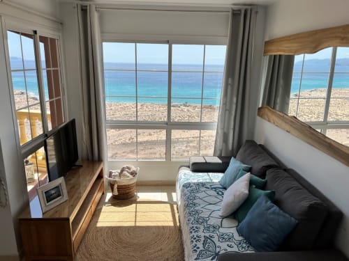 Sala de estar de apartamento frente al mar con vistas al océano, Smart TV, sofá negro y mar turquesa más allá de la playa arenosa